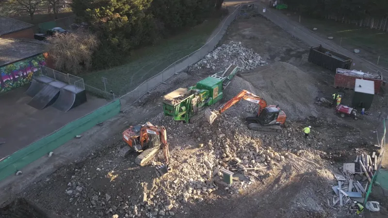 Aerial view of a demolition site with a green mobile crusher, two Hodge excavators working the rubble pile and workers in hi-vis