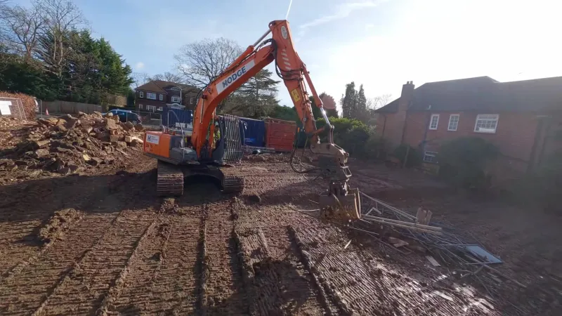 Hodge excavator with a sorting grab picking up metal debris on a cleared house plot, a pile of rubble behind it
