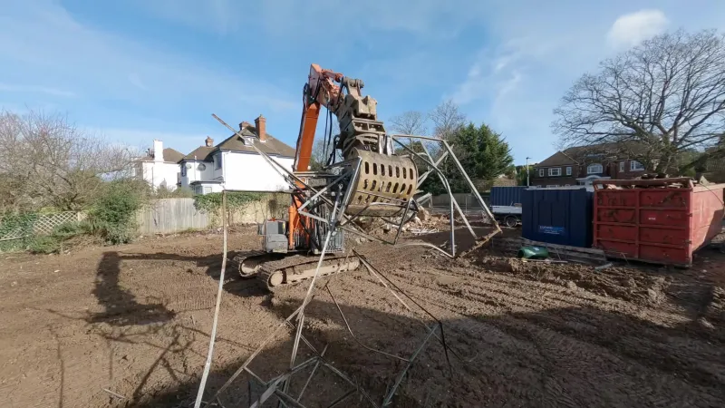 Excavator with a sorting grab lifting a tangle of metal framing on a cleared plot, with site containers and a skip to the right