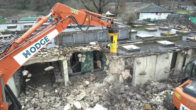 Hodge excavator with a hydraulic breaker demolishing a single-storey concrete building, seen from a drone