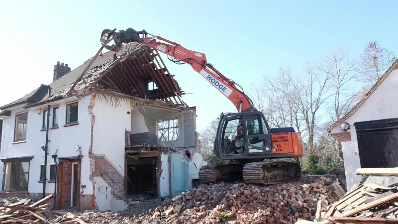 Hodge excavator with a demolition grab pulling the roof off a two-storey house, one side already open to the rooms