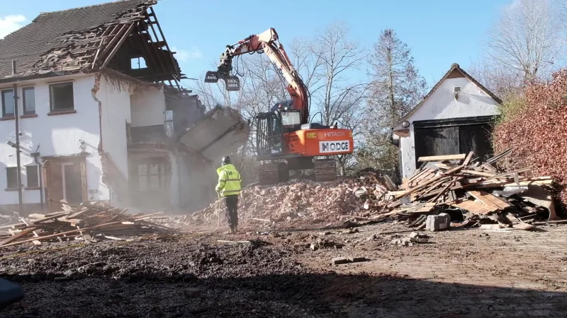 A worker in hi-vis, seen from behind, holding a hose in the dust while a Hodge excavator pulls down the rest of a house