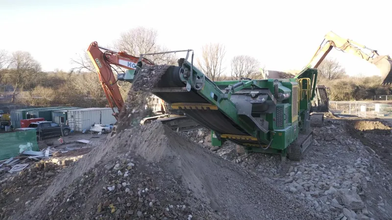 Mobile crusher dropping crushed demolition rubble onto a stockpile, with two excavators behind it