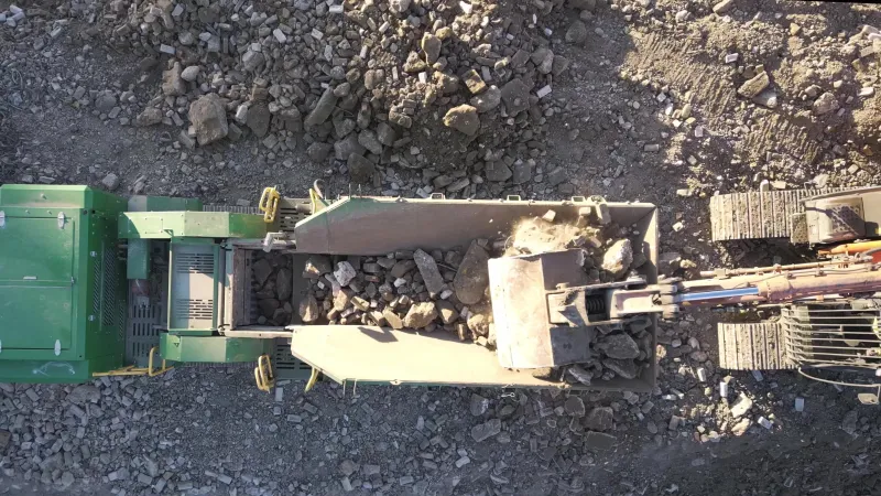 Overhead view of an excavator bucket tipping broken concrete and brick into the hopper of a green mobile crusher